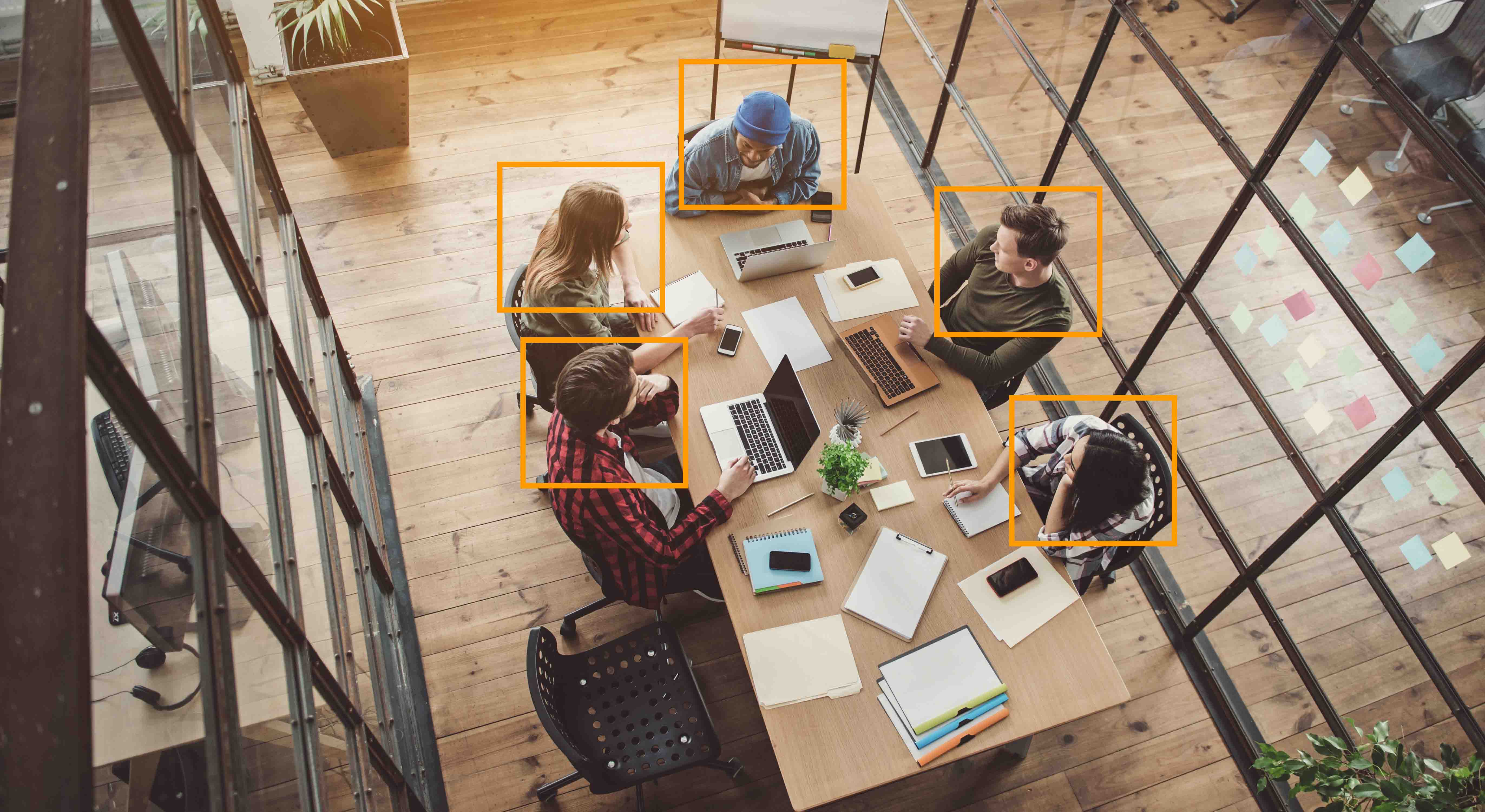 People sitting at conference table with laptops and phones. Orange squares are over their heads.