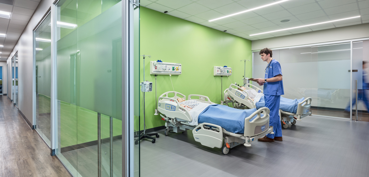 A nursing student stands at a simulated patient's bedside