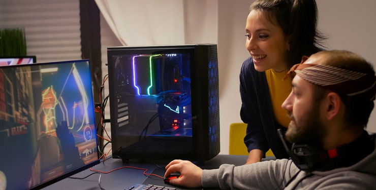 Two people sit at a desk and play a game on a computer in front of them