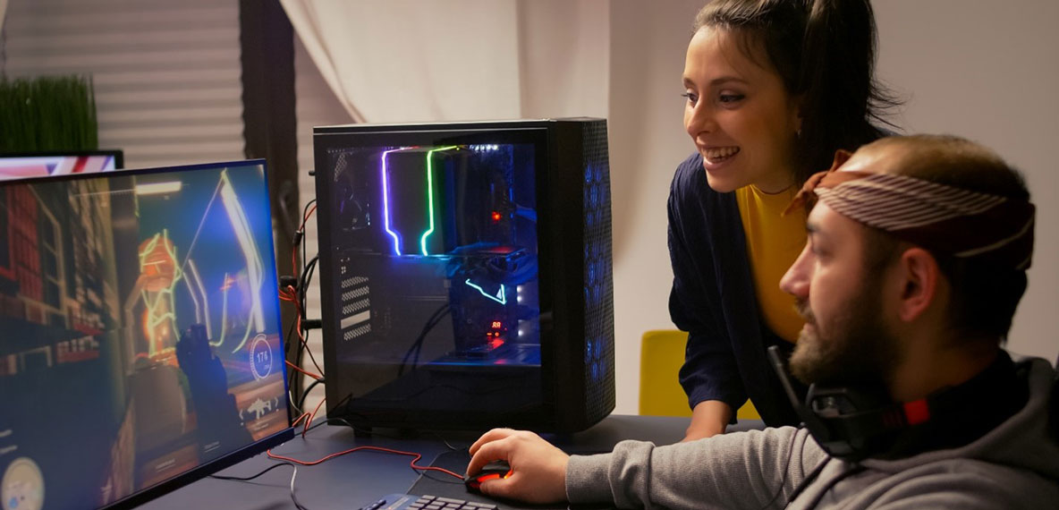 Two people sit at a desk and play a game on a computer in front of them