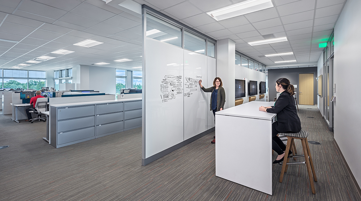 A collaboration zone—equipped with white boards and monitors—is near employee workstations.