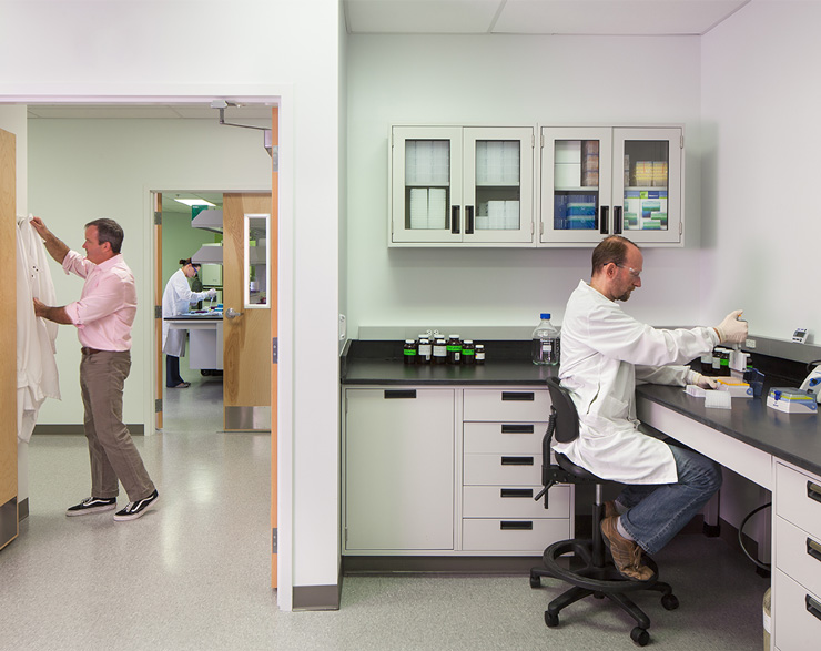 One man enters the lab, another is sitting down and using test tubes