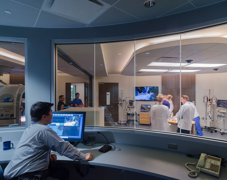 A man sits inside a room with a glass window looking into a lab full of students