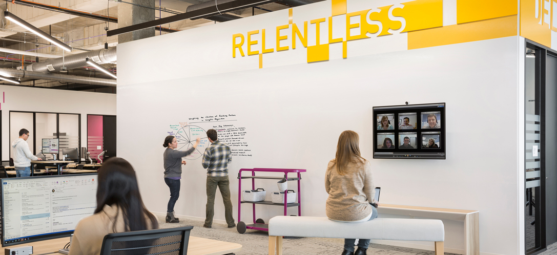 Inside an office, employees write on a nearby wall and use a screen to host a virtual meeting