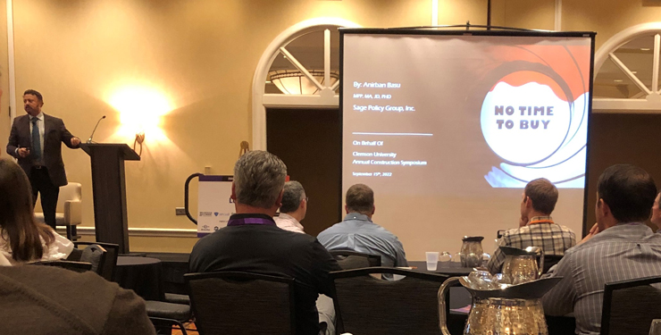 A speaker presents at a podium with a slide show to his right