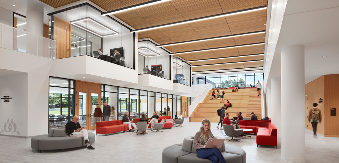 Two-story campus atrium with students studying on red and gray lounge seating below, wood-clad tiered stair seating, and glass-walled study rooms visible on the upper level.