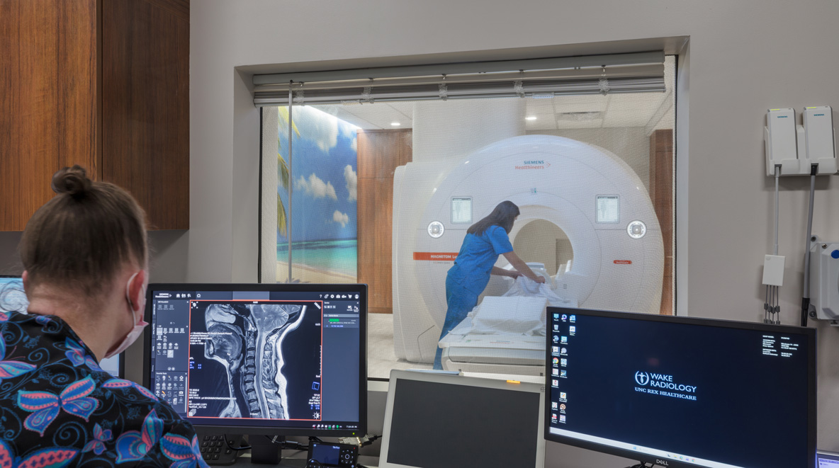  A technician monitors an MRI scan on a computer screen in the control room while another staff member prepares a patient on the scanner table in the adjoining room, visible through a window.