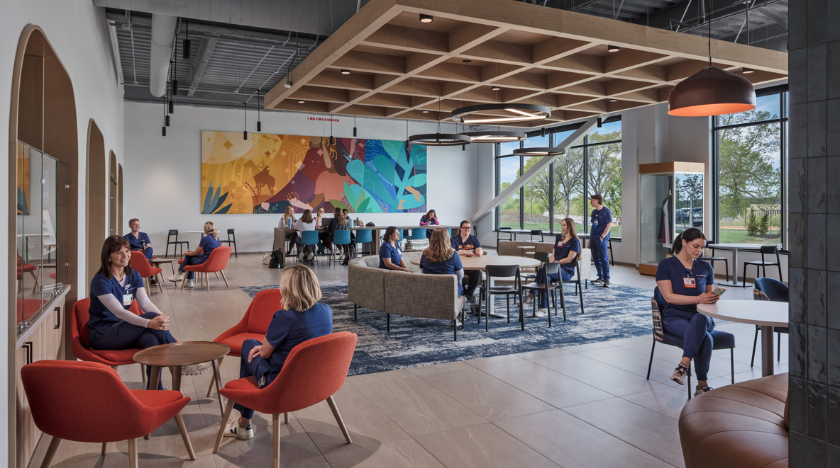 Healthcare staff in scrubs relaxing and chatting in a bright break room with orange lounge chairs, wood-grid ceiling detail, and a colorful mural along the back wall.