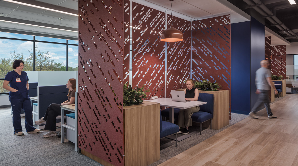 Open office workspace with a woman working on a laptop in a booth-style nook framed by perforated red decorative panels, plus workstations and a hallway visible alongside.