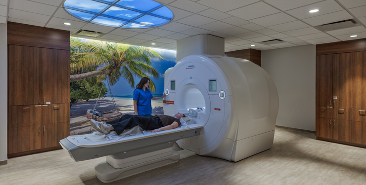 a look into the new MRI suite where the machine is in the middle of the room, there is a calming image on the far wall of a beach, and a new skylight feature on the ceiling projecting sky views