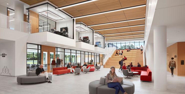 Two-story campus atrium with students studying on red and gray lounge seating below, wood-clad tiered stair seating, and glass-walled study rooms visible on the upper level.