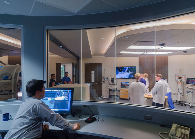 A man sits inside a room with a glass window looking into a lab full of students