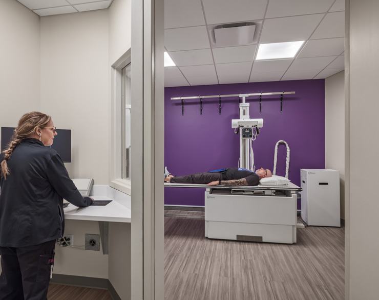 a look into an x-ray imaging room in the urgent care with a patient on the bed in the far part of the space and a x-ray tech in a viewing room on the left