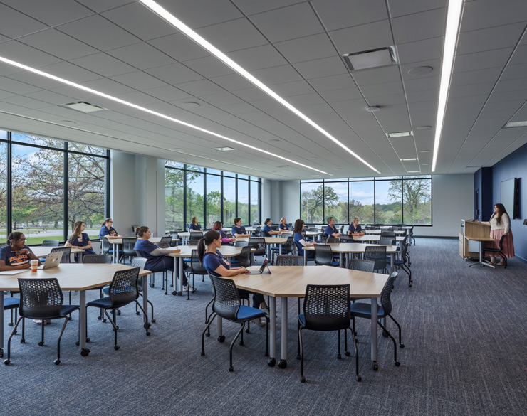 Bright classroom with students at group tables while an instructor leads a lesson from the front, floor-to-ceiling windows along one side.