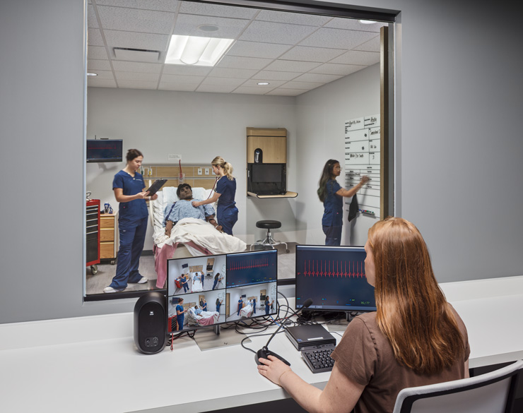 Control room monitoring a nursing simulation, with a technician watching camera and vital-sign feeds while staff practice patient care in the room beyond.
