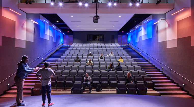 Two people stand on stage in a theater with audience members watching from their seats