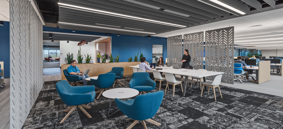 Collaborative hub at Forcepoint's Austin, TX office with a patterned dark ceiling grid, decorative screen dividers, and teal lounge chairs surrounding a communal work table.
