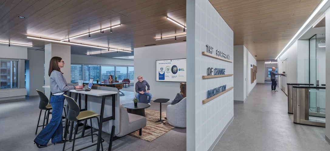Modern office corridor at P&G Canada's headquarters with a wood-slat ceiling, a standing-height meeting area with high-top seating, and a feature wall reading messaging about innovation near a turnstile entrance.