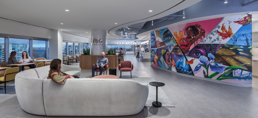 Lounge area at P&G Canada's headquarters with a curved gray sofa, city-view windows, and a large colorful mural featuring portraits and nature imagery along the corridor wall.