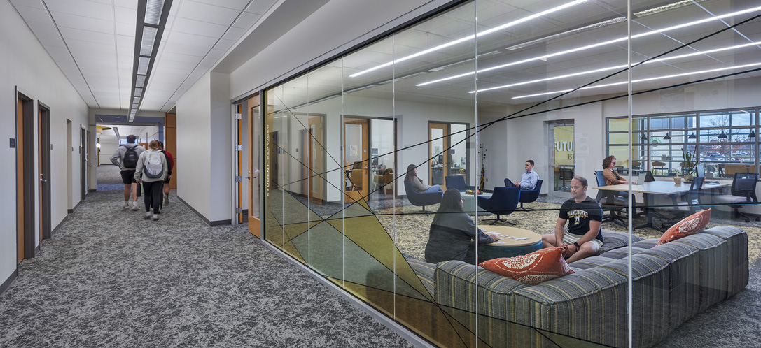 Glass-walled student services lounge at Manchester University's Fort Wayne campus, with a striped sectional couch, seating areas, and workstations visible through floor-to-ceiling windows along a hallway.