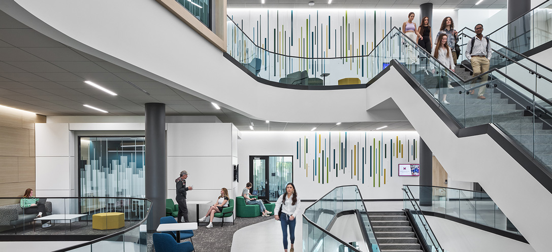 Multi-level atrium at Miami University's McVey Data Science Center with a sweeping staircase, colorful lounge seating below, and a striped vertical art installation spanning the wall.
