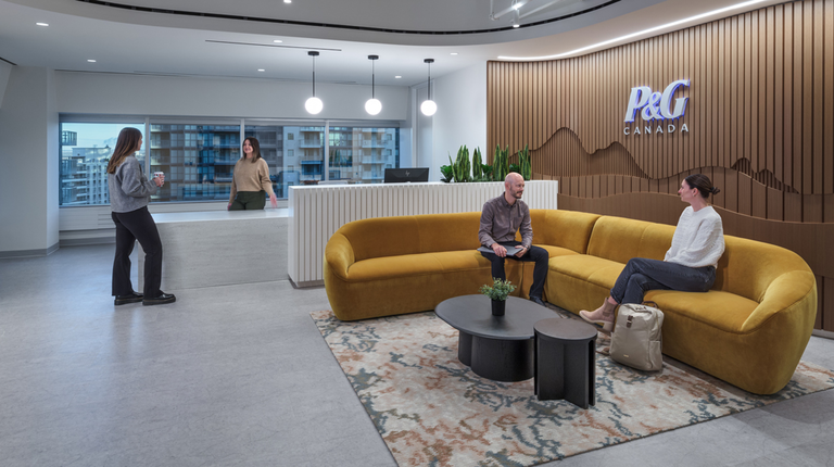 Employees sit on a brown velvet couch which is backed by a wall that says "P&G Canada"
