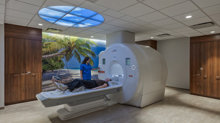 MRI room designed with calming beach decor, including a tropical mural and sky ceiling panel, while a technician assists a patient on the scanner table.