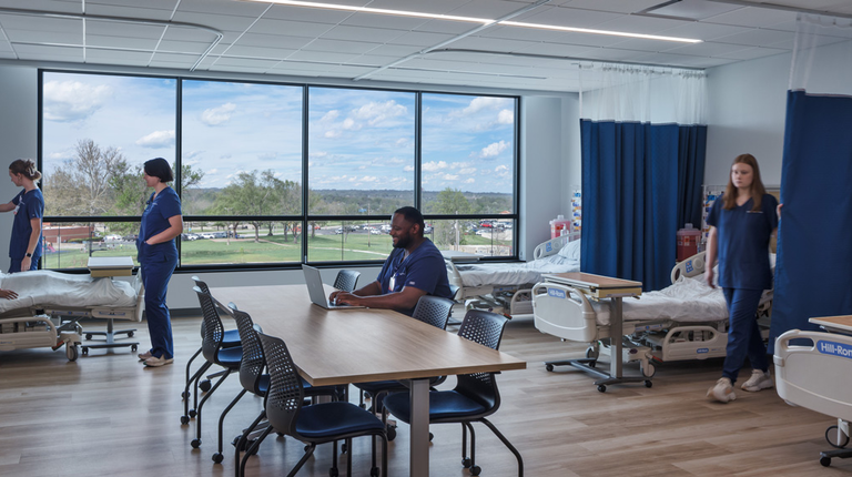 Nursing simulation lab with patient beds, staff in scrubs, and a central work table by large windows.