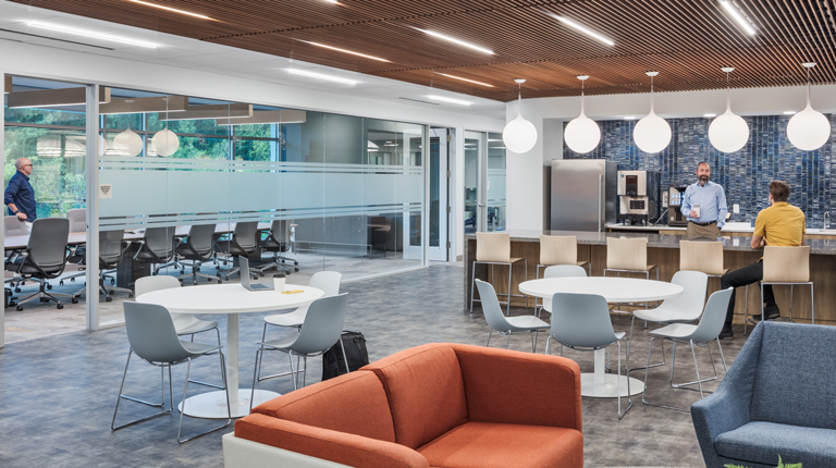 Two employees talk in their office kitchen while another works in a conference room